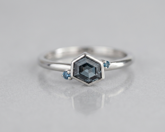 Geometric 9ct white gold stacking ring with an asymmetrical hexagon-cut grey-blue spinel and two blue diamond accents, part of the Geo range. Shot against a soft grey background.