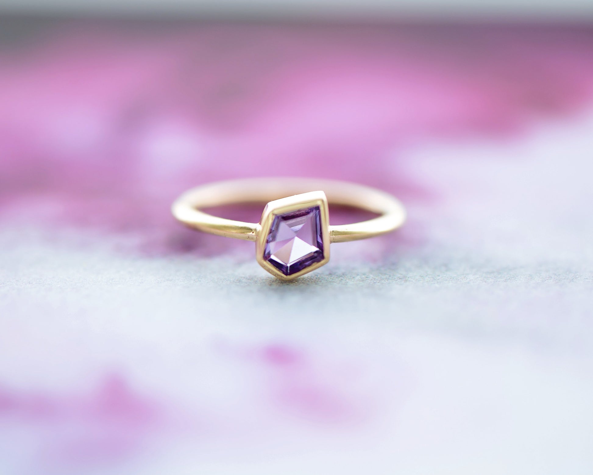 Asymmetrical geometric 9ct yellow gold stacking ring with a 0.72ct natural pink sapphire Geo cut center stone in a protective bezel setting, photographed on a soft beige felt surface with a subtle reflection. Part of the stackable Geo Range.