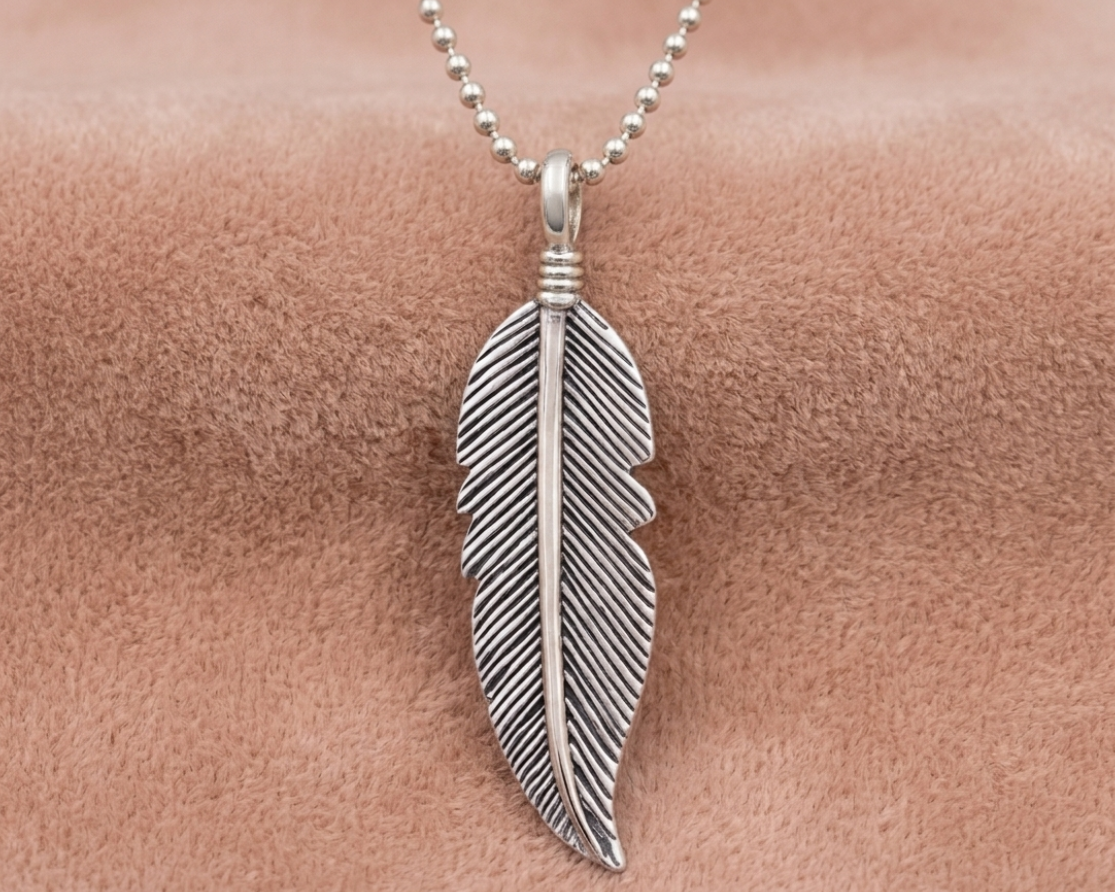 A detailed sterling silver feather pendant with an antique finish to highlight the texture shown on a neutral velvet background.