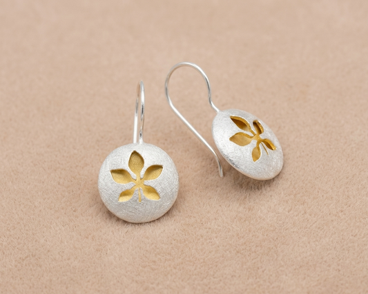 Handcrafted sterling silver heart stud earrings with a textured organic finish and a central yellow gold plated accent resting on soft skin-tone velvet.